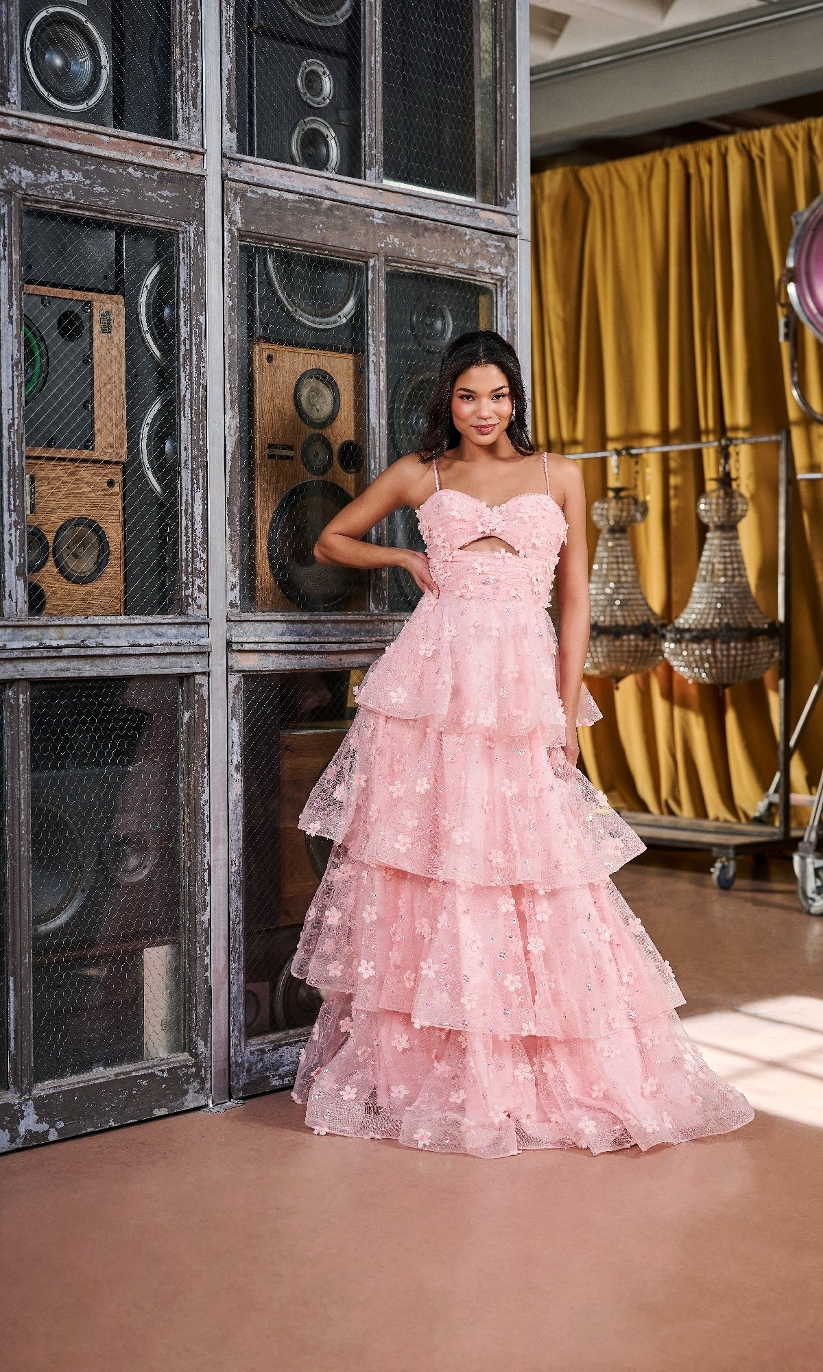 A woman stands indoors before metal speakers, wearing the Tiffany 16144 long prom dress, featuring a pink sweetheart neckline, thin straps, front cutout, and ruffled A-line skirt. Chandeliers and yellow curtains decorate the background.